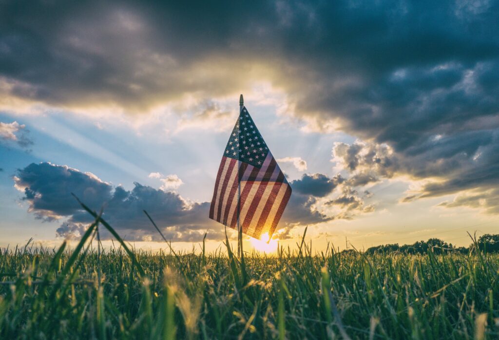 An American flag is seen against backdrop of grass and sky with sun and clouds.